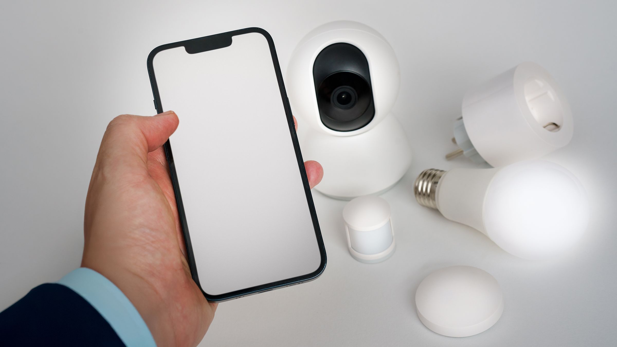 A smartphone with a blank screen held beside a home camera, smart plug, sensor and light bulb